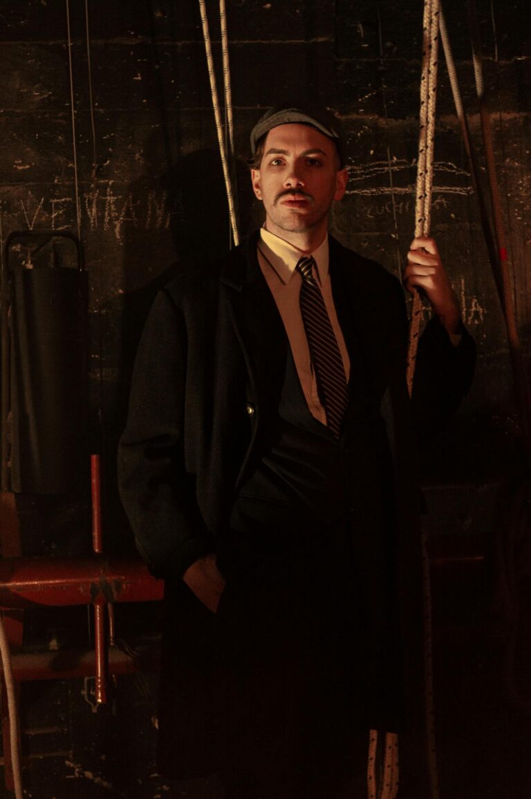A man in a suit and hat stands in a dimly lit vintage setting with ropes and shadows.